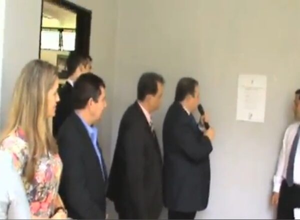 Foto da inauguração da sede do CONEF em Brasília. 4 pessoas aparecem olhando para uma placa fixada na parede, que está ao fundo: um homem de terno e gravata com o microfone na mão, outro em frente a ele com camisa e gravata e calça social preta, outros 2 de terno e gravata e uma mulher loira de vestido florido.