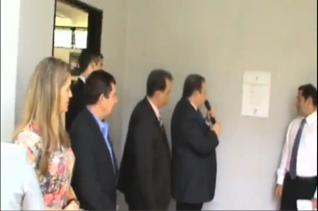 Foto da inauguração da sede do CONEF em Brasília. 4 pessoas aparecem olhando para uma placa fixada na parede, que está ao fundo: um homem de terno e gravata com o microfone na mão, outro em frente a ele com camisa e gravata e calça social preta, outros 2 de terno e gravata e uma mulher loira de vestido florido.