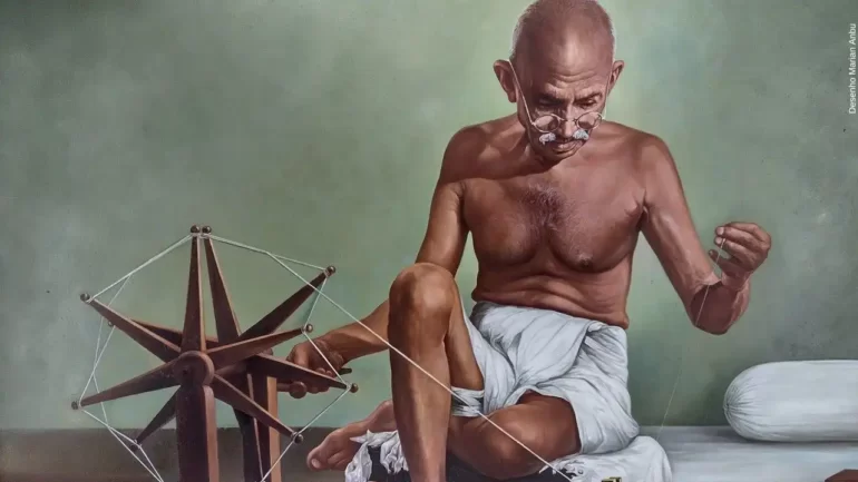 desenho do Maratma Gandhi sentado ao chão, vestindo apenas uma espécie de calção de pano de algodão branco, usando óculos de grau redondo e utilizando o tear.