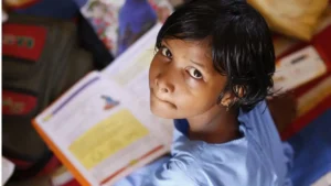 foto de cima para baixo de uma menina estudantes morena, de uns 12 anos, com cabelos lisos pretos, olhos pretos, com um caderno escolar aberto a frente, olhando para cima na direção da câmera. Usa blusa azul. Ao fundo a imagem desfocada de móveis e brinquedos.