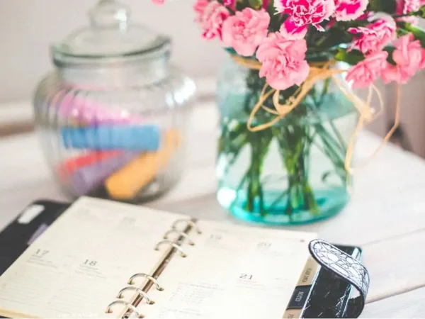 Foto de uma agenda de couro preta de planejamento diário, um vaso de vidro com 3 flores rosas e um pote de vidro com tampa com marcadores de textos dentro na cor laranja, azul, rosa, vermelho, lilás. Todos esses itens em cima de uma mesa de madeira.