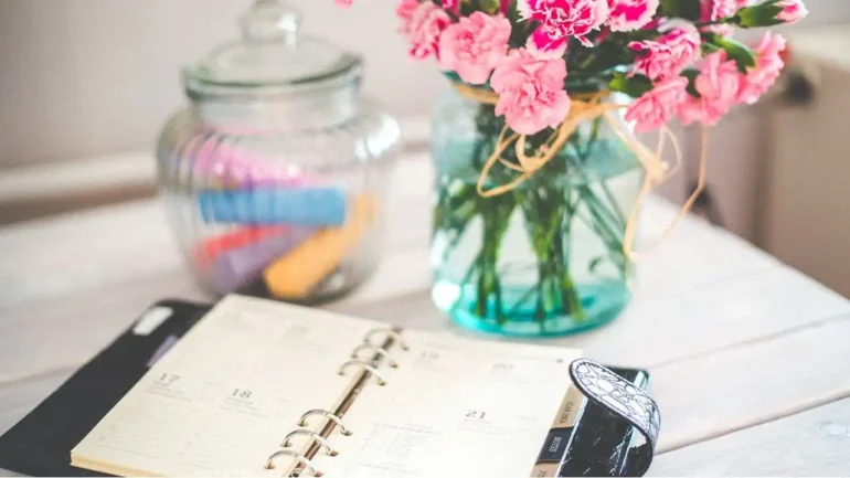 Foto de uma agenda de couro preta de planejamento diário, um vaso de vidro com 3 flores rosas e um pote de vidro com tampa com marcadores de textos dentro na cor laranja, azul, rosa, vermelho, lilás. Todos esses itens em cima de uma mesa de madeira.