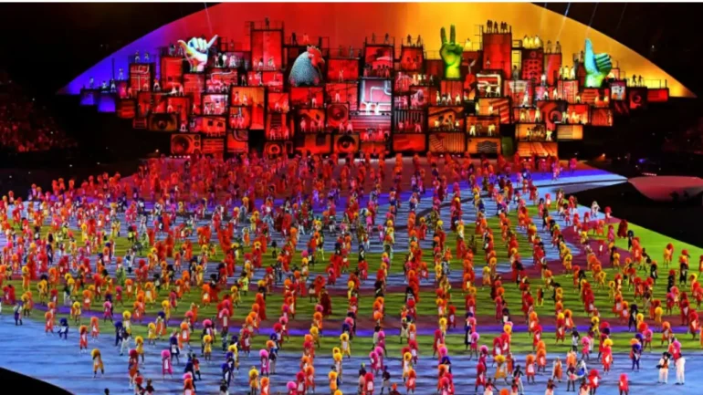 foto da abertura das Olimpíadas da Rio 2016, ao fundo um cenário colorido com muitas cores das favelas brasileiras e no centro do palco do estádio Maracanã centenas de pessoas em roupas coloridas dançando. Tudo com muitas luzes coloridas dão um espetáculo onde a cor laranja se destaca.