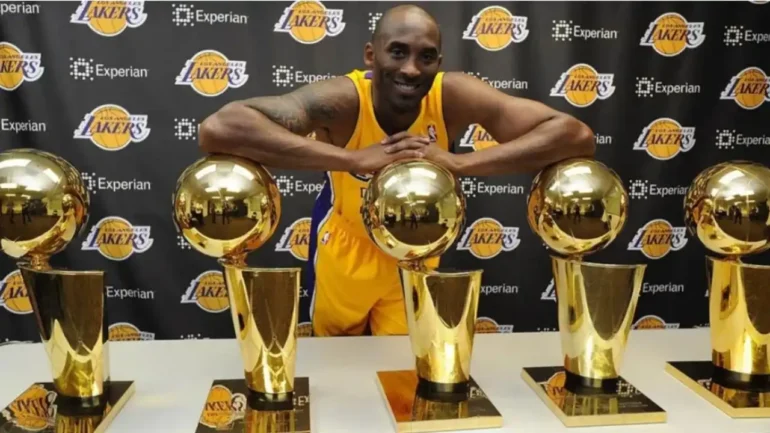 Foto do pentacampeão de basquete pela NBL Kobe Bryant vestido com o uniforme do LAKERS com 5 troféus a sua frente em cima de uma mesa e ele um pouco inclinado apoiando os cotovelos em 2 deles e sorrindo. Ao fundo um banner com várias logomarcas da equipe Lakers.