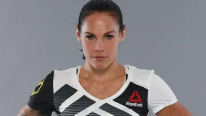 foto da bonita lutadora de MMA norte americana Lina com cabelos escuros em rabo de cavalo, vestindo blusa branca da marca REEBOK com detalhes pretos com as mãos na cintura.