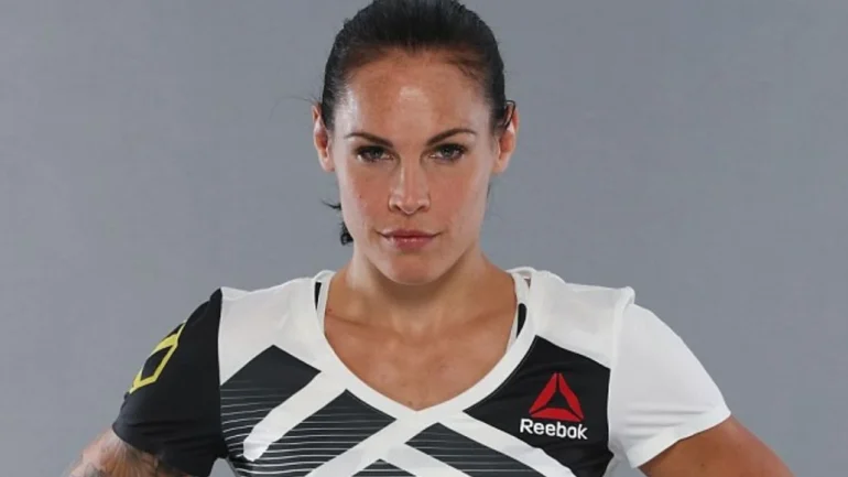 foto da bonita lutadora de MMA norte americana Lina com cabelos escuros em rabo de cavalo, vestindo blusa branca da marca REEBOK com detalhes pretos com as mãos na cintura.