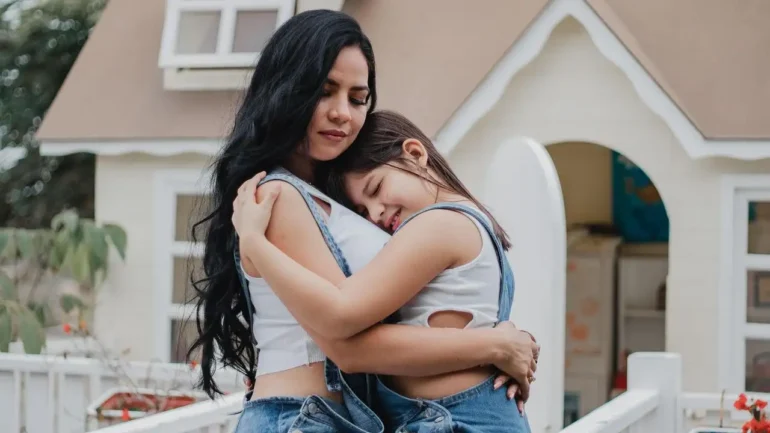 foto de uma moça branca cabelos longos pretos, vestindo camiseta em mangas branca e calças jeans abraçando uma menina vestindo macacão jeans e camiseta branca. As duas com os olhos fechados. Ao fundo a lateral de uma bela casa cor bege com janelas e detalhes brancos, em um dia claro.