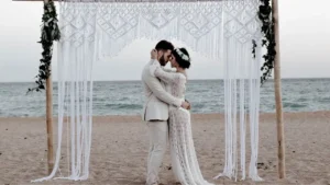 foto de um casal de noivos abraçado olhando um para o outro em ritual de casamento na areia da praia. Ele veste terno branco e usa baraba preta e cabelos pretos, ela vestido branco de renda transparente, cabelos castanhos curtos e coroa na cabeça.