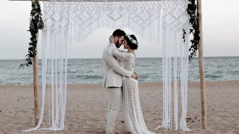 foto de um casal de noivos abraçado olhando um para o outro em ritual de casamento na areia da praia. Ele veste terno branco e usa baraba preta e cabelos pretos, ela vestido branco de renda transparente, cabelos castanhos curtos e coroa na cabeça.