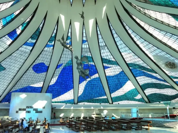 foto do teto da Catedral de Brasília, uma obra do arquiteto Niemeyer, com ladrilhos azul e branco. Com 16 colunas brancas em forma de bumerangue e com uma cinta de concreto que se unem no topo e 3 estátuas de anjos pinduradas. Em primeiro plano, uma jovem de calça jeans e camiseta olha para as imagens.