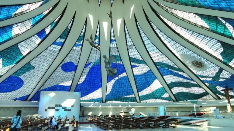 foto do teto da Catedral de Brasília, uma obra do arquiteto Niemeyer, com ladrilhos azul e branco. Com 16 colunas brancas em forma de bumerangue e com uma cinta de concreto que se unem no topo e 3 estátuas de anjos pinduradas. Em primeiro plano, uma jovem de calça jeans e camiseta olha para as imagens.