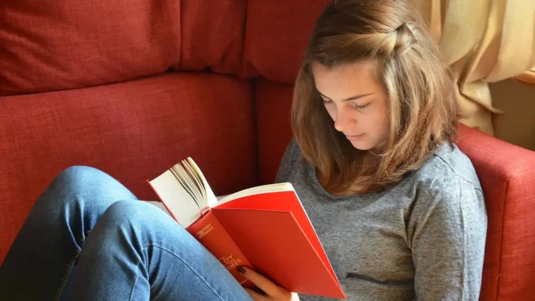 foto de uma bonita moça loira sentada em um sofá vermelho escuro, com os cabelos soltos e com um grampo ao lado segurando a franja, vestindo blusa cinza e calaças jeans elndo um livro de capa vermelha.