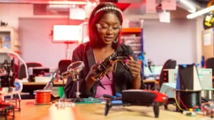 foto de uma mulher negra, com rosto de alegre, usando óculos de proteção transparente, em um laboratório bem colorido de robótica. Ela esta segurando uma espécie de drone desmontado. Há outros em cima da sua mesa e muitas luzes, estantes atrás e várias ferramentas e materiais.