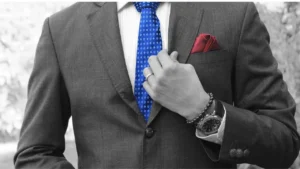 imagem em preto e branco do tronco de um homem vestindo paletó e gravada, sendo que a gravata é azul e o lenço no bolso é vermelho. Ele usa uma pulseira de couro e um relógio esportivo e segura a lapela do paletó com a mão esquerda, a do relógio.