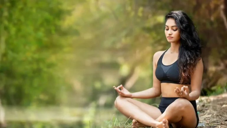 foto da uma mulher morena clara com cabelos castanhos escuros soltos, vestindo suquini preto de 2 peças, sentada no chão de areia, com as pernas cruzadas e as mãos sobre os joelhos, em posição de meditação e com os olhos fechados. Ao fundo, a natureza e a iluminação do dia.