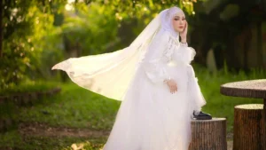 foto de uma mulher morena clara com turbante e véu e vestido de noiva branco, com o pé esquerdo em cima da um banco de pedras, calçando um sapato preto fechado com salto baixo, com a mão esquerda no queixo olhando para a câmera. Ao fundo, gramas e árvores.