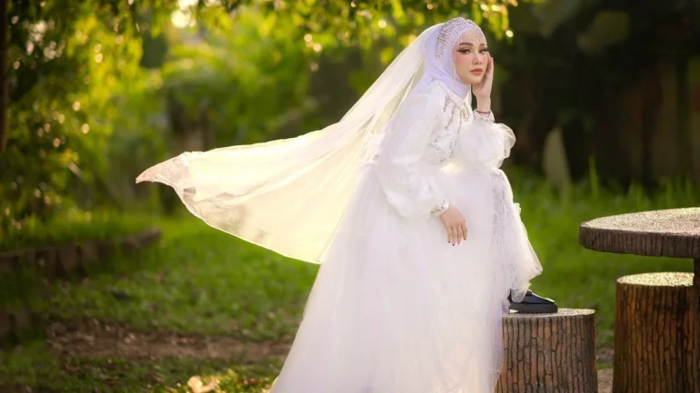 foto de uma mulher morena clara com turbante e véu e vestido de noiva branco, com o pé esquerdo em cima da um banco de pedras, calçando um sapato preto fechado com salto baixo, com a mão esquerda no queixo olhando para a câmera. Ao fundo, gramas e árvores.