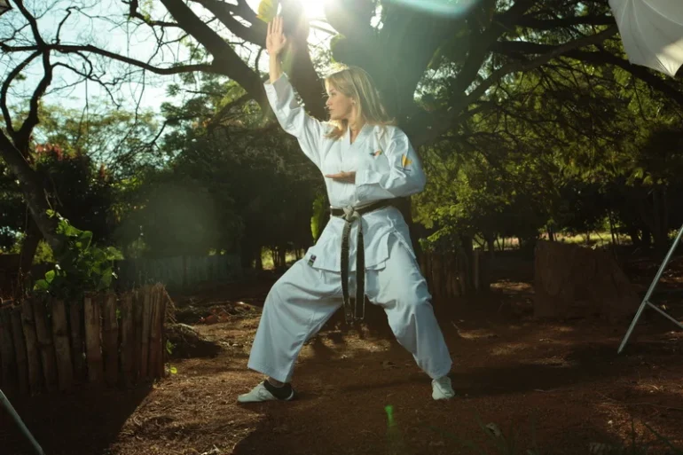 foto da carateca loira Carla Ribeiro, com os cabelos soltos, vestindo quimono de karate branco, faixa preta, fazendo uma base kokutsu do kankudai. Ao fundo &aacute;rvores do cerrado, e ch&atilde;o de terra.