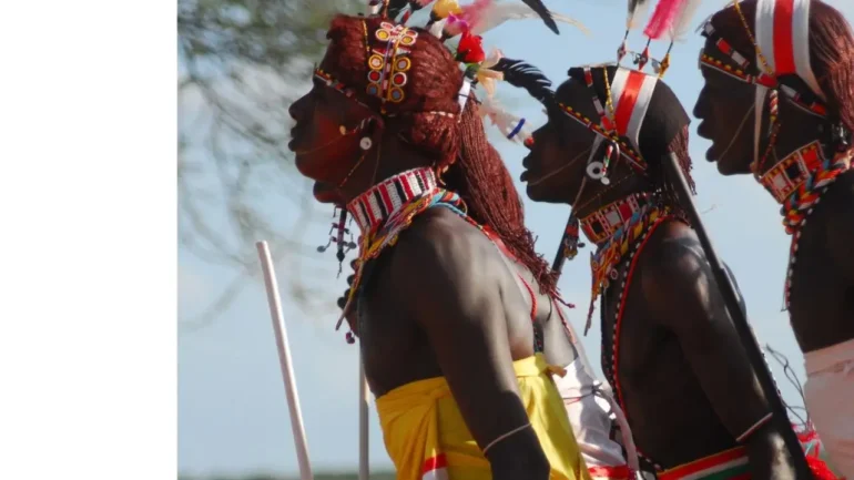 Foto de lado de quatro guerreiros negros, com vestidos bem coloridos, colares coloridos, brincos coloridos, segurando lanças em gestos de um ritual. Ao fundo o céu azul claro do dia e uma árvore.