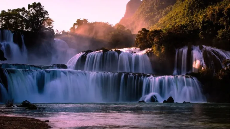 foto de uma cascata de pequenas e belas cachoeiras. Ao fundo, a luz solar batendo nas montanhas.