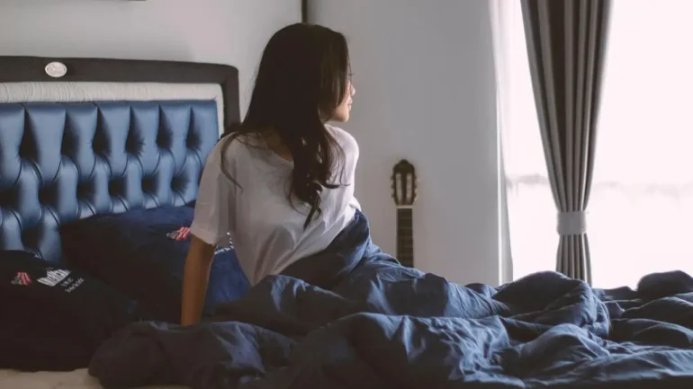 Mulher de cabelos castanhos escuros, vestindo camisola branca, sentada na cama, de baixa do edredon azul, olhando um pouco para a janela, onde entra a claridade da manhã, e tem uma cortina branca transparente e um cortinado cinza amarrado. A cama tem cabeceira azul almofadada, na mesma cor do edredon. O quarto tem paredes brancas e tem um violão encostado na parede, que fica no mesmo lado da janela.