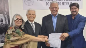 foto de 4 pessoas segurando um contrato e olhando para a câmera, uma senhora de cabelos brancos vestindo uma chale, um senhor de cabeça branca vestindo terno completo azul marinho, um senhor de cabelos brancos vestindo terno com camisa azul sem gravata, um senhor de cabelos pretos vestindo terno azul royal e camisa preta. Todos sorridentes.