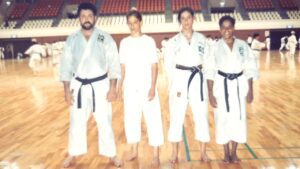 Foto da equipe feminina de karate e do seu técnico. Da esquerda para a direita: o técnico Antônio Testa, vestindo quimono branco de karate e faixa preta; a atleta Carla Ribeiro usando calça de karate e camiseta branca; a atleta Simone Nicola usando quimono e faixa preta; e a atleta Maria Cecília de Almeida, a Ciça Maia, usando quimono e faixa preta. Dentro do ginásio de esporte de Fukuoka, no Japão. O Chão do ginásio é de madeira clara envernizada bem bonito.