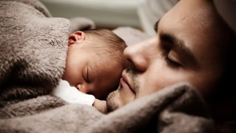 um pai deitado com os olhos fechados e o fillho bebezinho deitado no seu peito dormindo, Os dois estão envoltos em um cobertor e a foto só mostra o rosto deles.