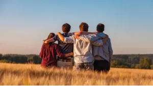 4 pessoas abraçadas de costas olhando para o horizonte a frente em um campo cheio de espigas para colher. DA esquerda para à direita: uma moça de camisa vermelha, um rapaz com blusa azul e calça bege com cinto, um rapaz com camisa de manga comprida azul clara quadriculada e uma calça quadriculada, e outro rapaz com agasalho de capuz cinza, uma camiseta cinza e uma calça azul com cinto marrom.