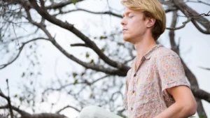 foto de um homem loiro, vestindo uma camiseta de botões estampada com clores claras, com os olhos fechados meditando. Ao fundo, vários galhos de uma árvore em um dia claro.