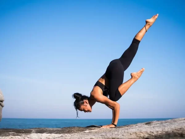 uma mulher vestida de top preto e calça de ginãstica preta apoiada sobre os 2 braços e com uma perna esquerda apoiada no cotovelo esquerdo e a outra perna esticada para cima. Ela está em cima de uma rocha e ao fundo se vê o céu azul. Uma posição típica de yoga.