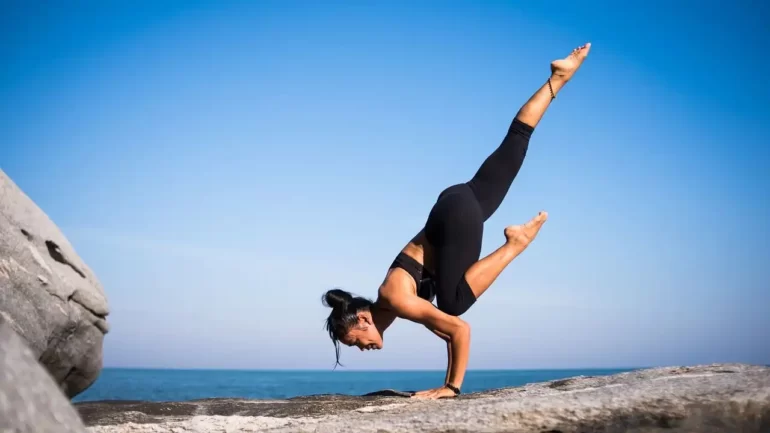 uma mulher vestida de top preto e calça de ginãstica preta apoiada sobre os 2 braços e com uma perna esquerda apoiada no cotovelo esquerdo e a outra perna esticada para cima. Ela está em cima de uma rocha e ao fundo se vê o céu azul. Uma posição típica de yoga.