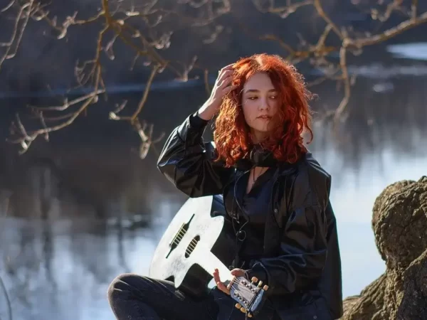 uma moça roiva de uns 20 anos, sentanda no chão, a beira de um rio, segugurando um violão com a mão esquerda e os cabelos com a direita.