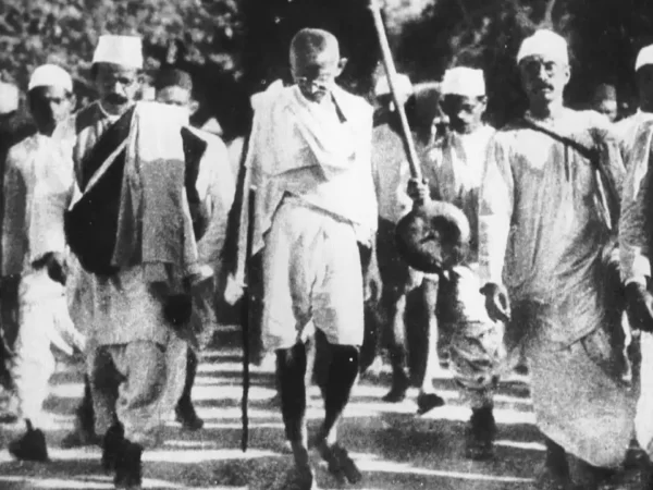 Foto em preto e branco com Maratma Gandhi andando cercado de uma multidão de seguidores, vestindo uma túnica branca e bermuda de algodão, óculos, sandálias e um cajado na mão.