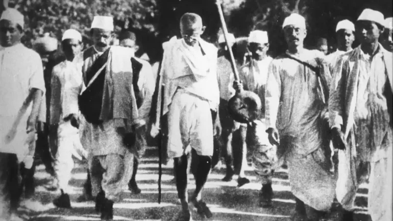 Foto em preto e branco com Maratma Gandhi andando cercado de uma multidão de seguidores, vestindo uma túnica branca e bermuda de algodão, óculos, sandálias e um cajado na mão.