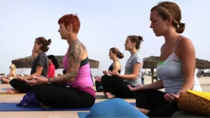 Na foto aparecem 11 mulheres sentadas em colchonetes coma as pernas cruzadas em posição meditativa, com os olhos fechados, na calçada de uma praia. No fundo aparece a areia, dois grandes sombreiros de palhas, e o mar ao longe, As mulheres vestem calças pretas de lycra e camisetas.