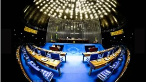 foto com a grande angular do plenário do Senado Federal em Brasília mostra as bancadas e poltronas da plenária e a mesa diretora a frente no palco. Ao centro no teto se destacam as luzes.