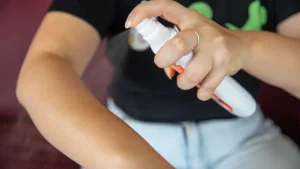 foto de parte do tronco de uma mulher com calça jeans e blusa preta segurando um repelente com a mão esquerda e asperjando o produto no braço direito que está levantado à sua frente.