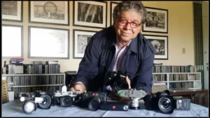 foto do fotógrafo Orlando Brito em seu apartamento, vestindo um casaco azul típico de fotógrafo e uma camisa social rosa clara e óculos vermelhos redondo segurando uma câmera fotográfica, apoiado em uma mesa com 6 outras câmeras com lentes a frente, Atrás, vários quadros emoldurados pendurados na parede.