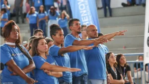 Foto de várias pessoas dentro de um ginásio vestindo camisetas azuis do Programa de capacitação do GDF, escritas: RENOVA DF. Elas estão com os braços direitos estendidos em posição de juramento. Em destaque, no primeiro plano, 5 pessoas: uma mulher negra, outra branca, uma mulata, um rapaz branco e um rapaz moreno. Ao fundo, uma bandeira com os dizeres RENOVA DF.