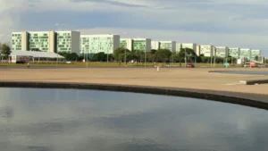 foto de 10 ministérios da capital do Brasil, em Brasília. Na frente, em primeiro plano parte de um espelho dágua cercado por cimento.