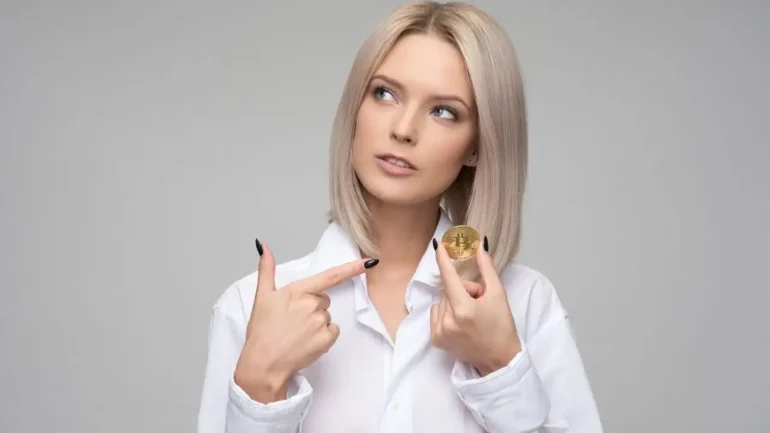 Foto de uma mulher loira, vestindo camisa branca, segurando uma moeda dourada com o símbolo da bitcoin e apontando para a moeda com a outra mão. Ela olha para cima à esquerda. O fundo é cinza claro.
