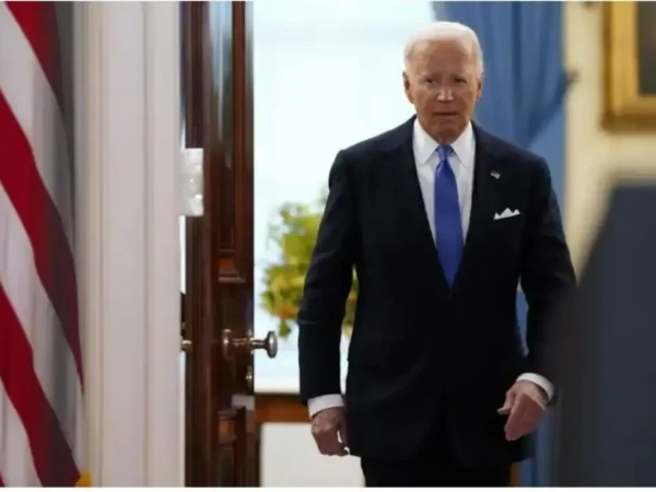 foto do presidente dos EUA Joe Biden saindo do seu gabinete, vestindo terno preto todo abotoado, blusa branca e gravata azul. A esquerda aparece parte da bandeira dos EUA, a direita parte de uma pintura em um moldura dourada.