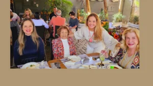 uma foto bem colorida com algumas pessoas sentadas em um restaurante. Em primeiro plano uma bonita jovem loira vestindo blusa azul, uma linda senhora vestindo blaser colorido e blusa branca, uma linda loira vestindo roupa bege e uma bonita senhora vestindo roupa estampada.