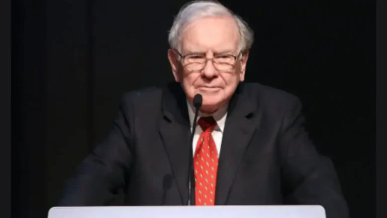 foto do mega empresário multi bilhonário Warren Buffet vestindo terno preto, camisa branca e gravata vermelha e óculos transparentes. Em um púlpito. O fundo é preto.