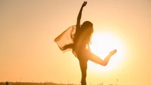 foto de uma mulher dançando com panos esvoaçantes, cabelos soltos, tendo o por do sol como fundo, o que permite ver mais o contorno do seu corpo.