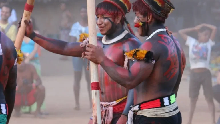 foto de 2 jovens indígenas vestindo calções, com os corpos pintados, usando colares e adereços de ritual típico. Ao fundo, é possível ver outros índios na aldeia.