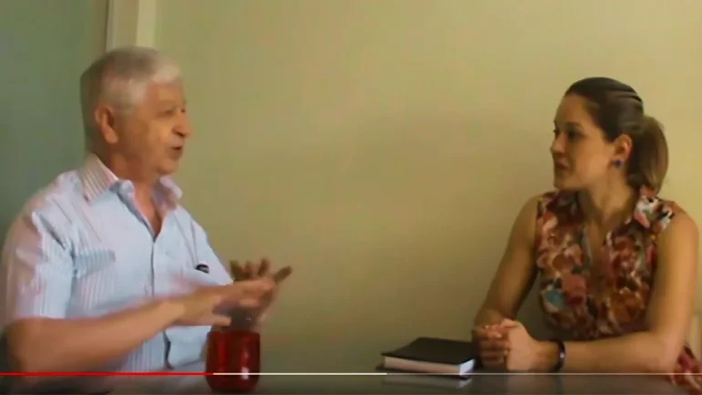 foto do Prof Pedro Demo vestindo camisa azul clara sem mangas, cabelos brancos sentado em conversa com Carla Ribeiro, vestindo vestido florido e cabelos loiros em rabo de cavalo. Os dois sentados em frente a uma mesa de vidro transparente.