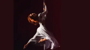 foto de uma mulher com vestido branco esvoaçante fazendo uma pose de dan;a pindurada em uma barra de Pole Dance. O fundo [e preto e os cabelos dela estão soltos, são da altura dos ombros e tem coloração ruiva.