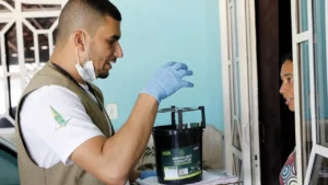 foto de um agente de vigilância sanitária falando com uma moradora na porta da casa dela, segurando uma prancheta e uma armadilha preta de plástico para mosquito aedes aegypt. Ele veste uma camiseta branca com logo do GDF na manga e um colete bege, além de uma máscara. Ela um vestido estampado.